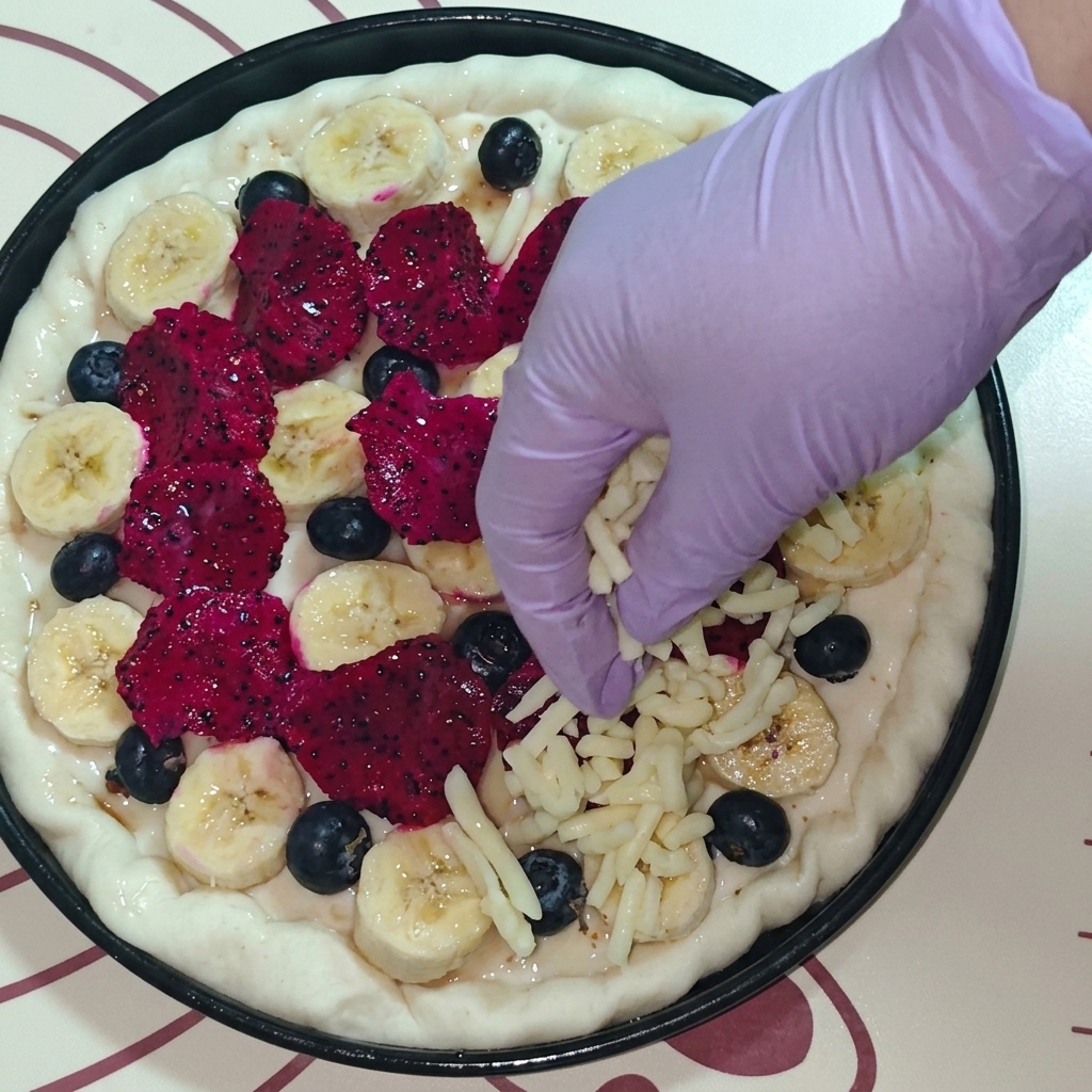 Vue de dessus dune main avec un gant violet saupoudrant de la mozzarella râpée sur une pizza garnie de fruits.