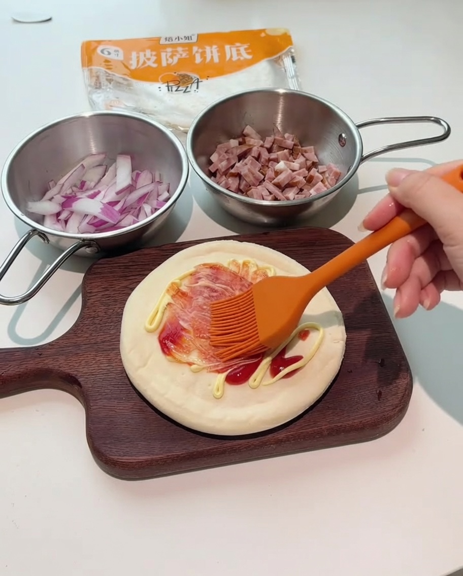 Un pinceau en silicone orange étalant les sauces rouge et blanche sur la base de la pizza.