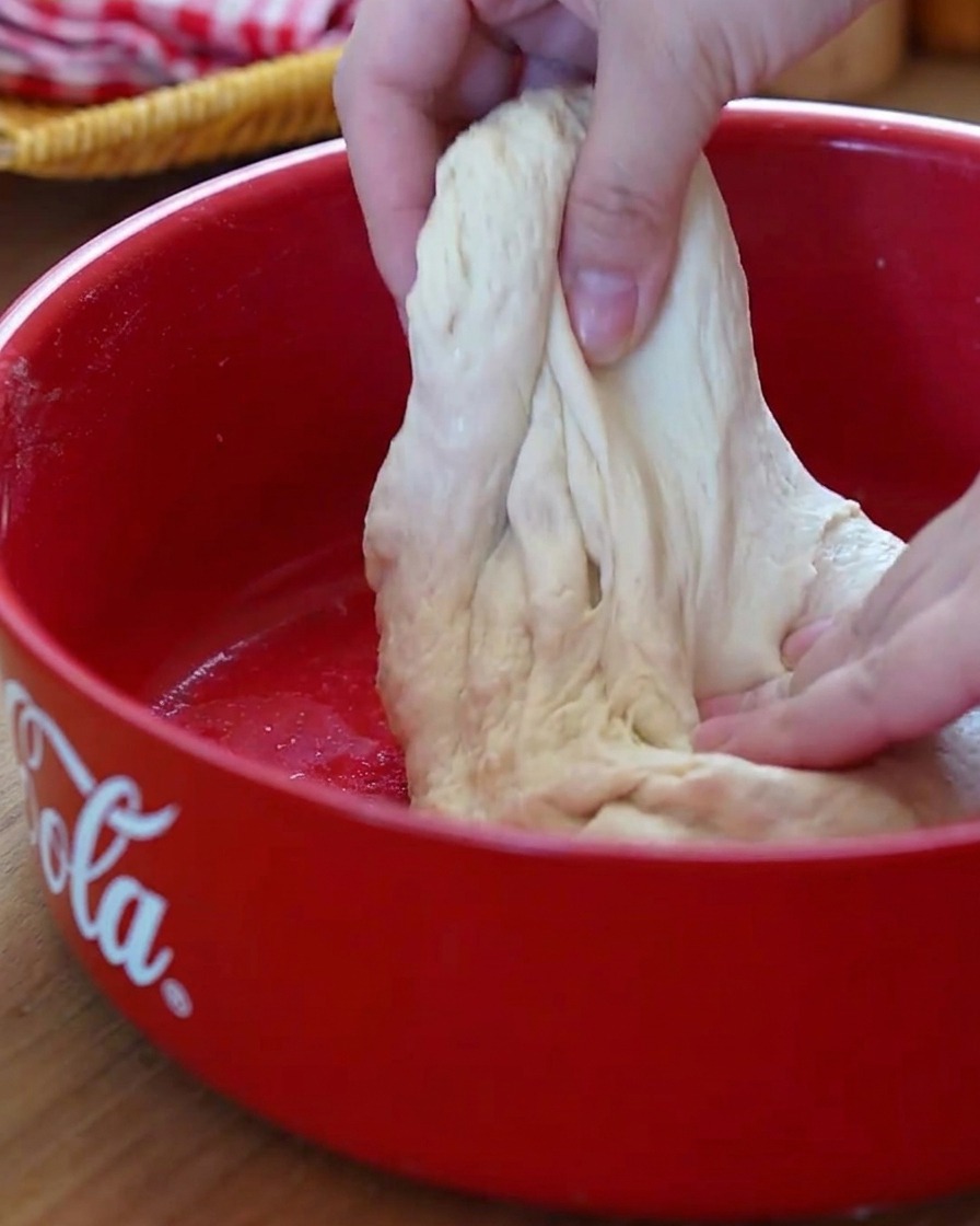 Gros plan sur des mains effectuant la technique du rabat sur une pâte à pizza souple dans un bol rouge.