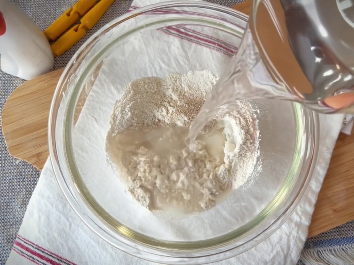 Un bol en verre contenant un mélange de farine, de levure, de sel et deau combiné avec une spatule.
