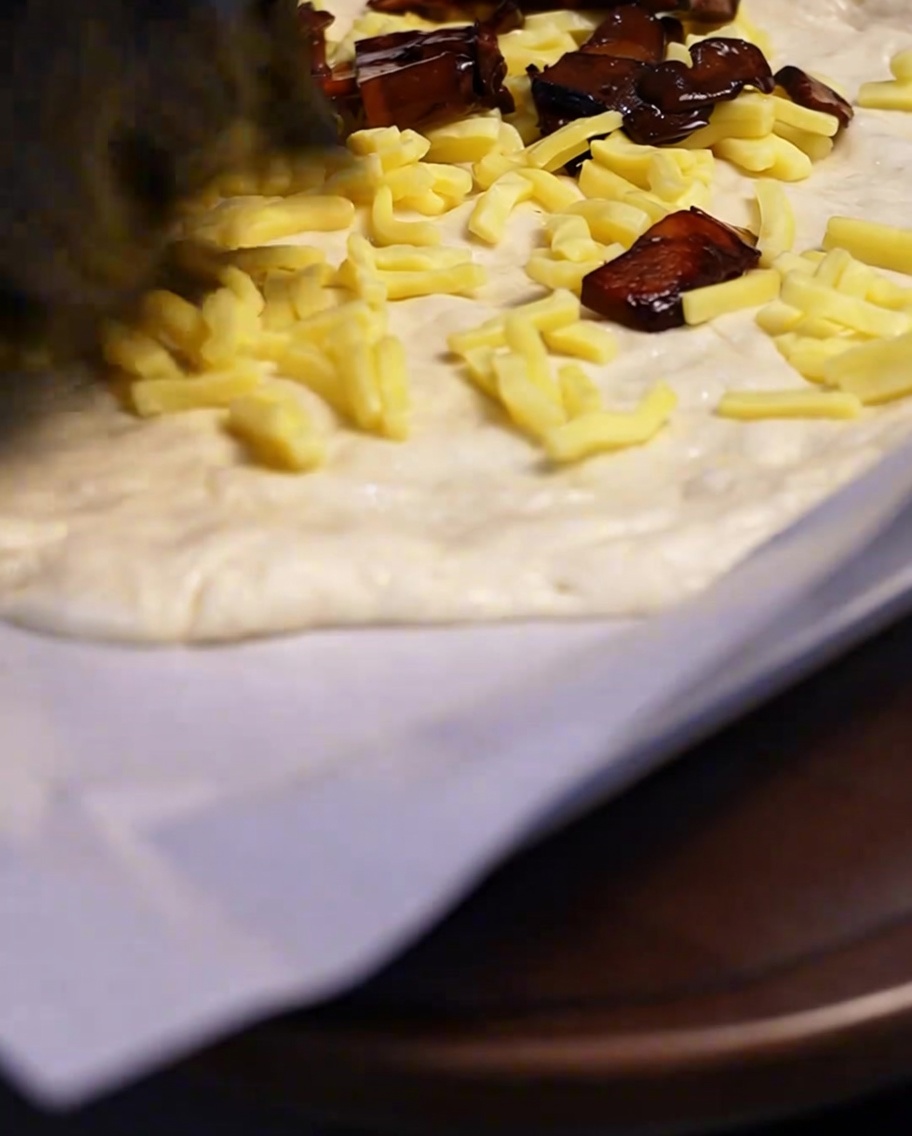 Des champignons cuits et assaisonnés déposés à la cuillère sur une couche de fromage râpé, sur une base de pâte à pizza.