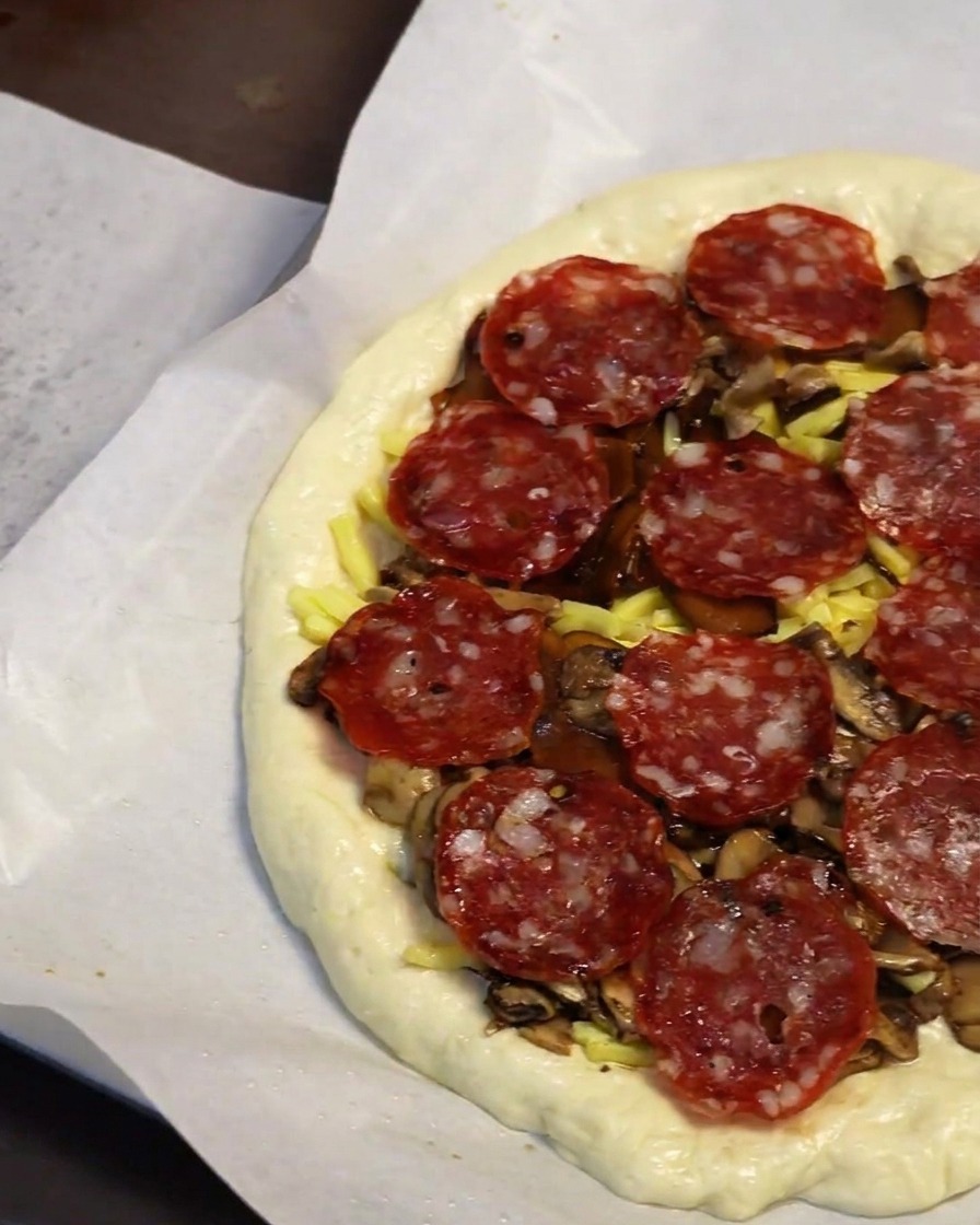 Gros plan dune main disposant des tranches rondes de pepperoni sur une couche de champignons et de fromage sur une pâte à pizza.