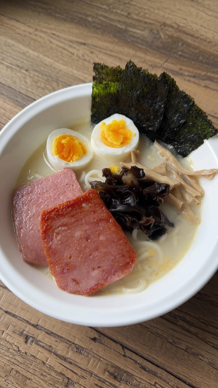 Un bol de ramen instantané garni de tranches de viande de déjeuner saisies, de champignons oreilles de bois terreux, de pousses de bambou et de nori croustillant.