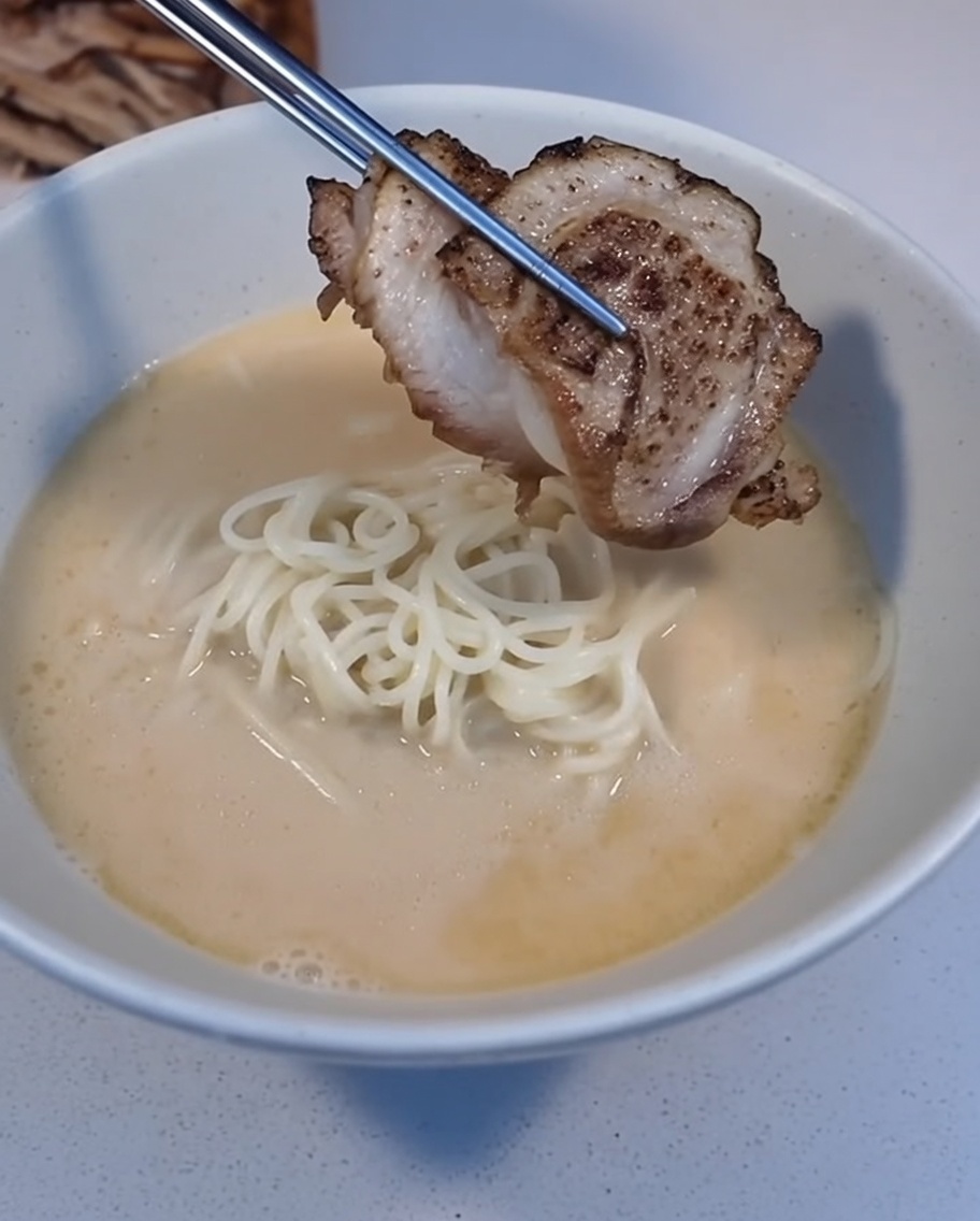 Une tranche de Char Siu braisée placée avec une pince sur un bol de ramen.