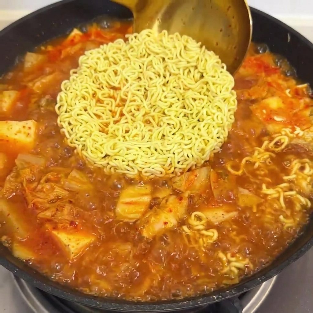 La sauce rouge versée dans une poêle avec des nouilles ramen bouillantes et le bouillon de kimchi.