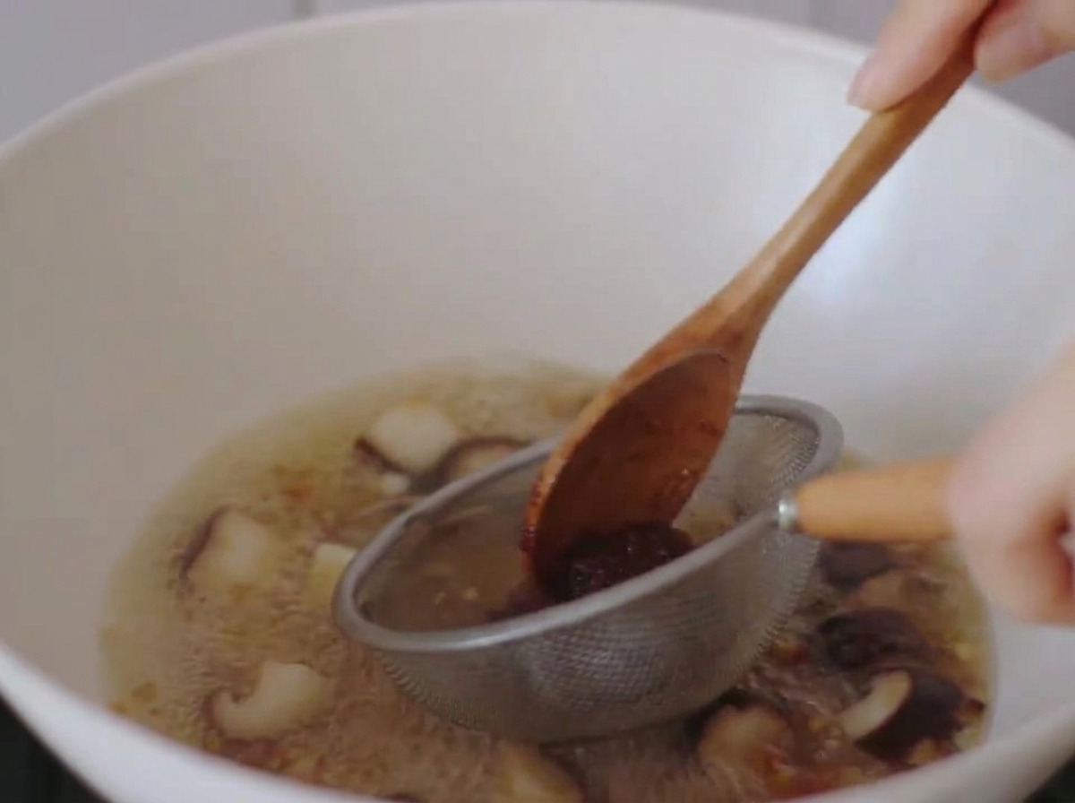 Pâte miso dissoute dans le bouillon bouillant à laide dune petite passoire métallique et dune cuillère en bois.