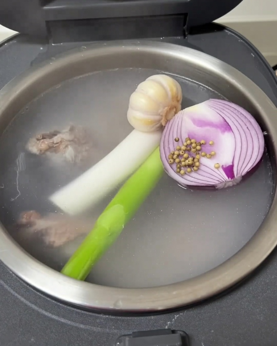 Vue intérieure du cuiseur à riz montrant lail, loignon, loignon vert et le poivre blanc flottant avec les travers de porc.