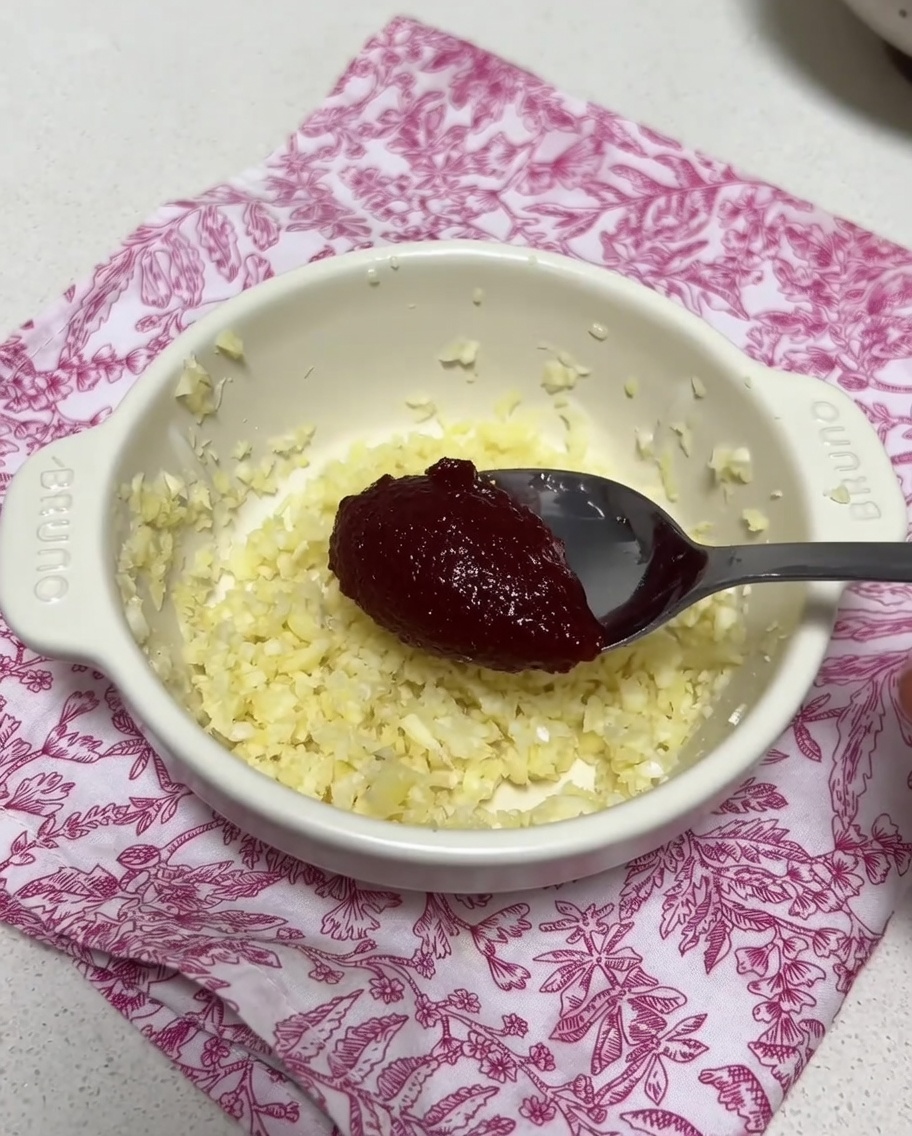 Un petit bol contenant de lail haché et une noisette de pâte de piment coréenne, mélangés avec une louche de bouillon.