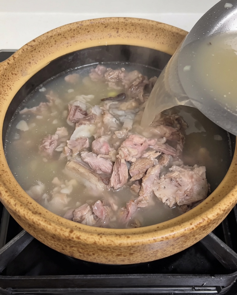 Un bouillon de viande chaud et opaque versé depuis un récipient en inox dans le pot en terre cuite contenant le porc et les pousses.
