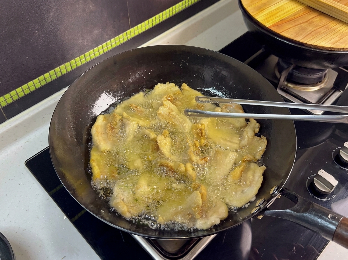 Tranches de poitrine de porc en train de frire dans un wok, retournées avec des pinces en métal.