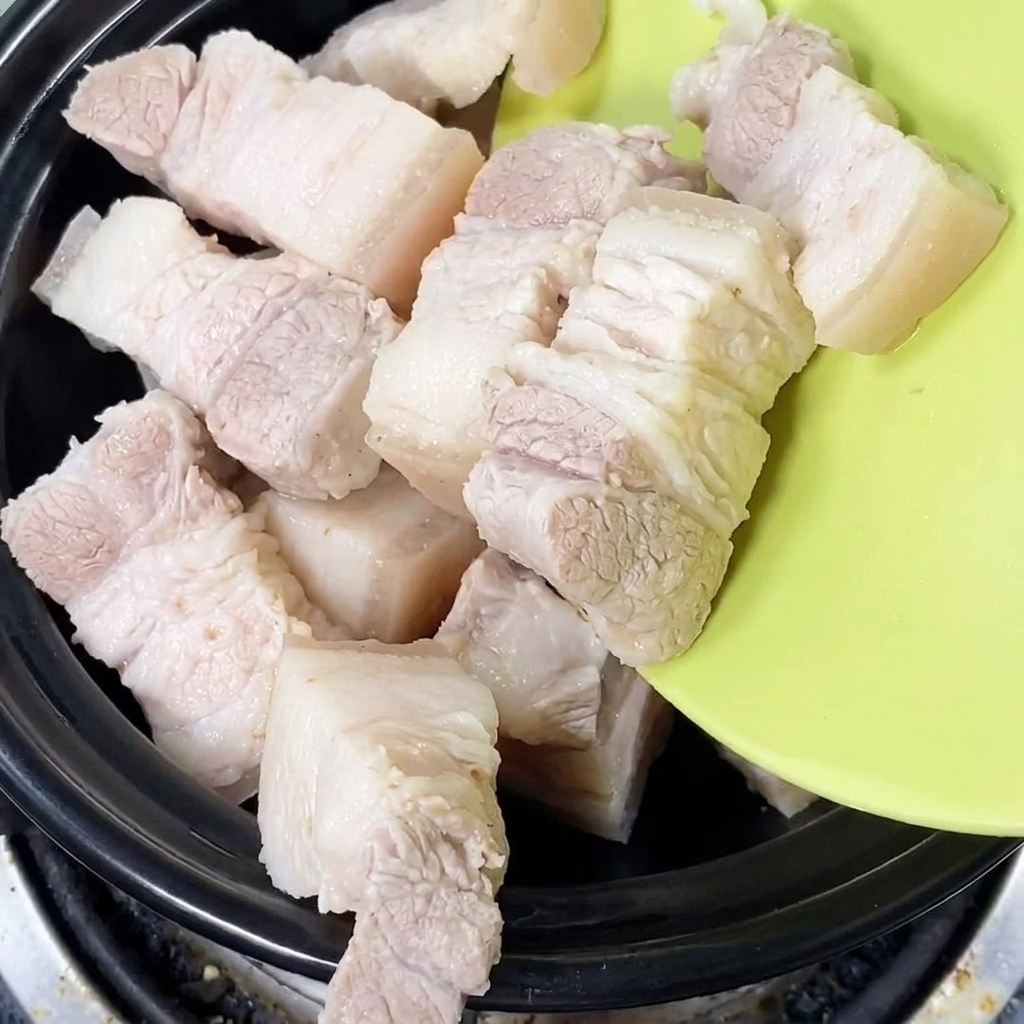 Morceaux de poitrine de porc fraîchement blanchis déposés dans une cocotte en terre cuite noire.