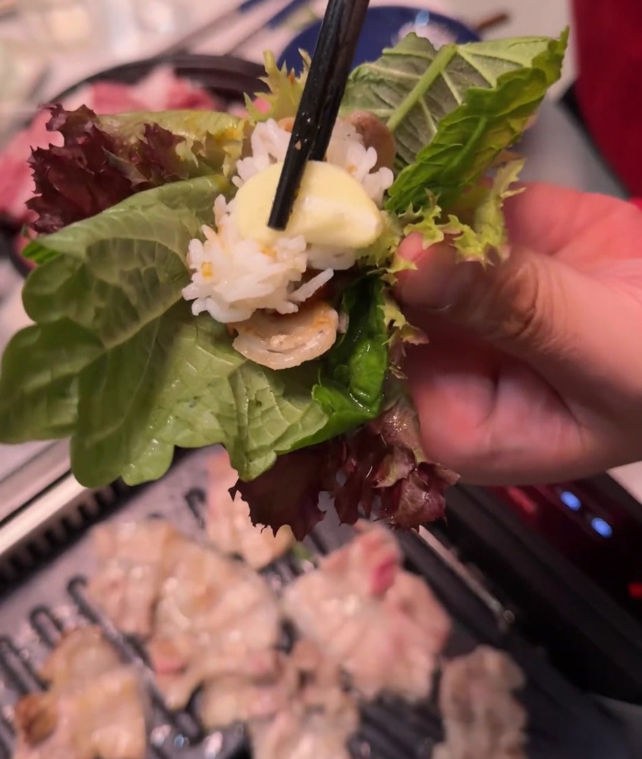 Une main tenant un wrap de laitue et perilla contenant viande, riz, pâte épicée et ail.