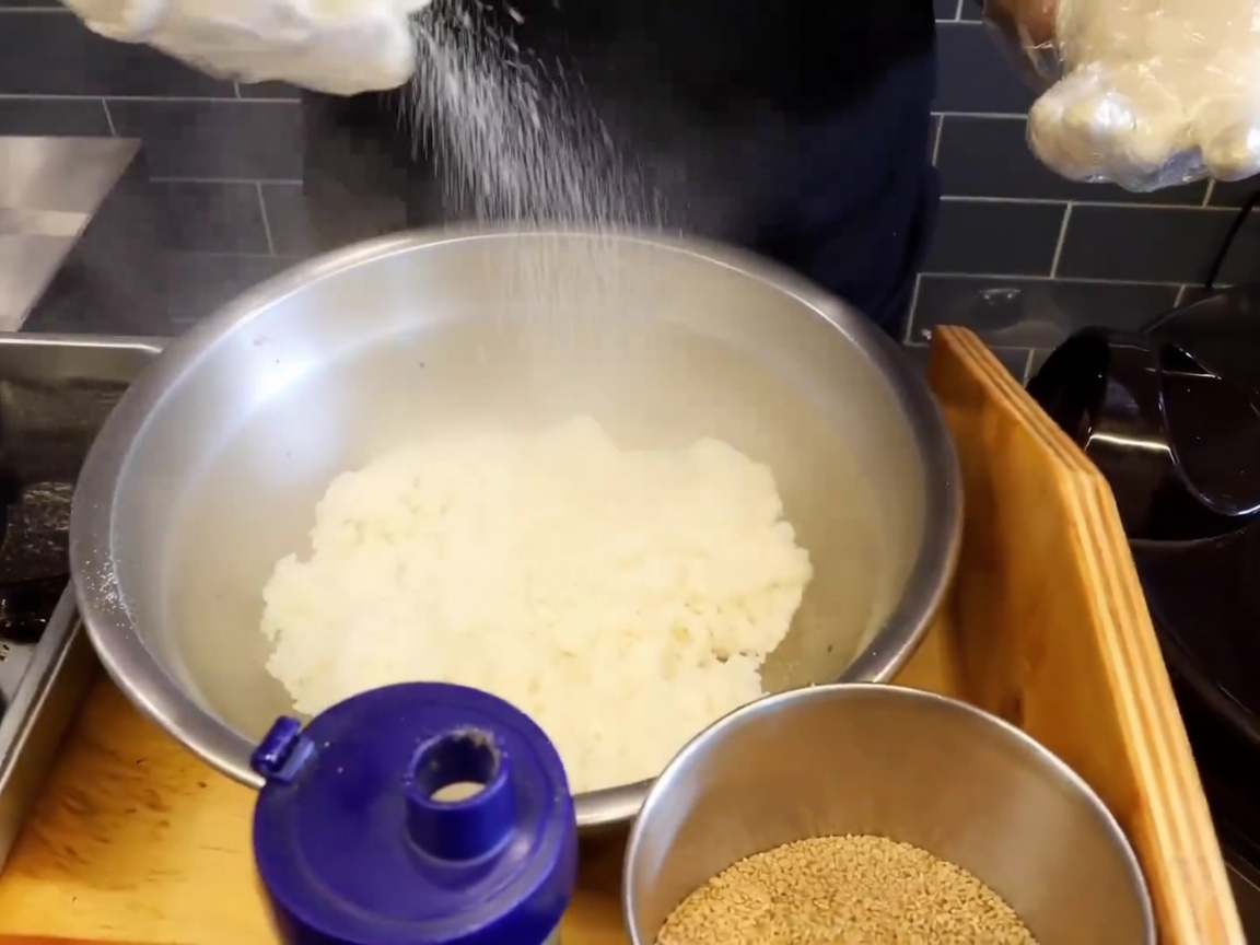 Gros plan sur un grand bol en inox rempli de riz gluant fumant, avec du sel et des graines de sésame saupoudrés sur le dessus.