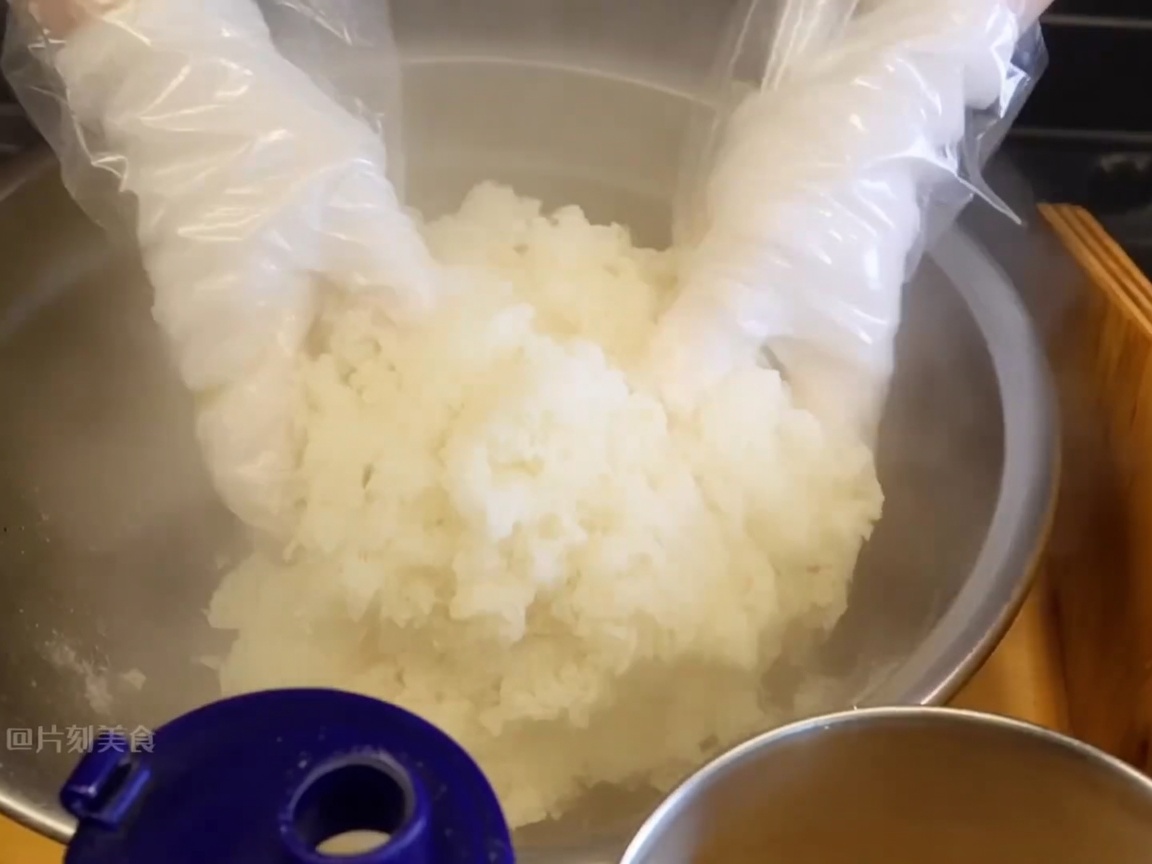 Mains gantées mélangeant du riz gluant fumant dans un grand bol pour incorporer les assaisonnements.