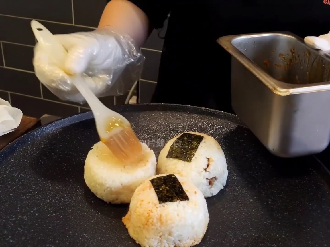 Boulettes de riz sur une plaque chaude, brossées avec un glaçage brun riche par une main gantée.