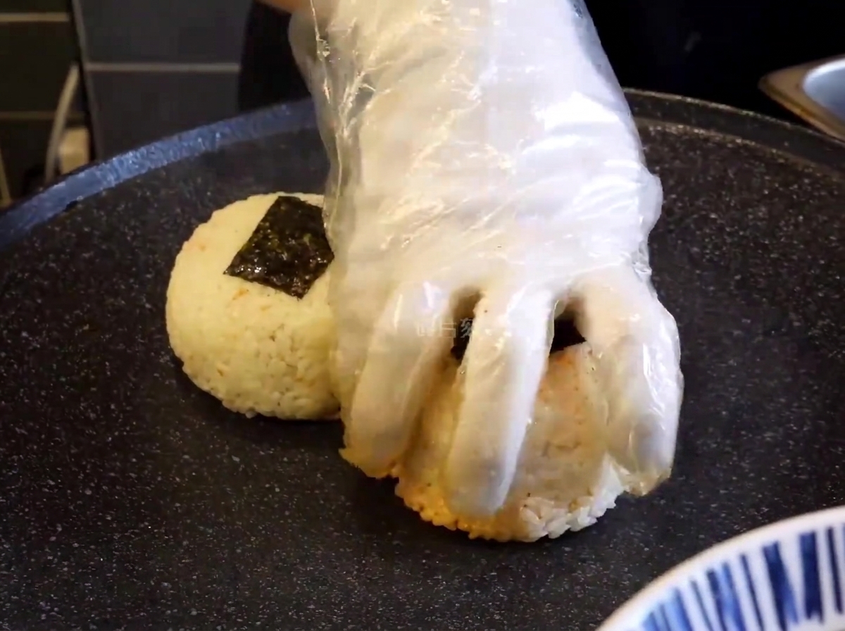 Gros plan sur une main gantée pressant des boulettes de riz sur une plaque noire chaude pour les saisir.