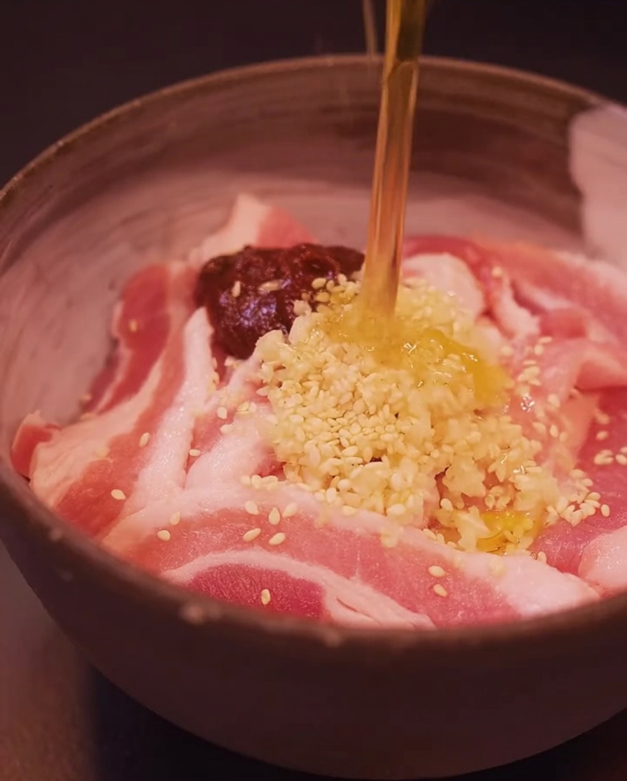 Tranches de poitrine de porc dans un bol, assaisonnées avec du gochujang, de lail, des graines de sésame et de lhuile de sésame.
