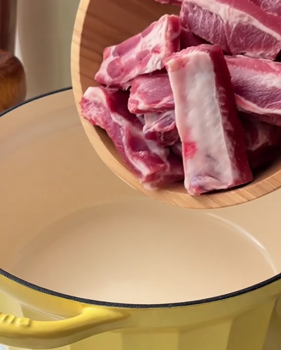 Morceaux de porc dans une cocotte jaune immergés dans leau avec des oignons verts et du gingembre, prêts à être blanchis.