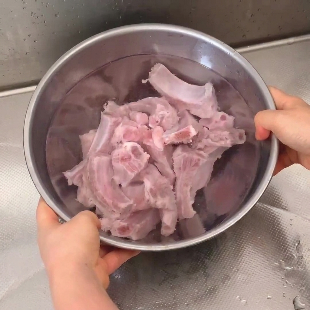 Travers de porc frais en train de tremper et dêtre rincés dans un bol en inox sous le robinet.