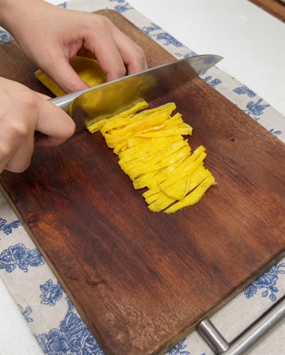 Vue rapprochée dune main utilisant un couteau de cuisine pour trancher une fine crêpe dœuf jaune cuite en lanières nettes sur une planche à découper en bois.
