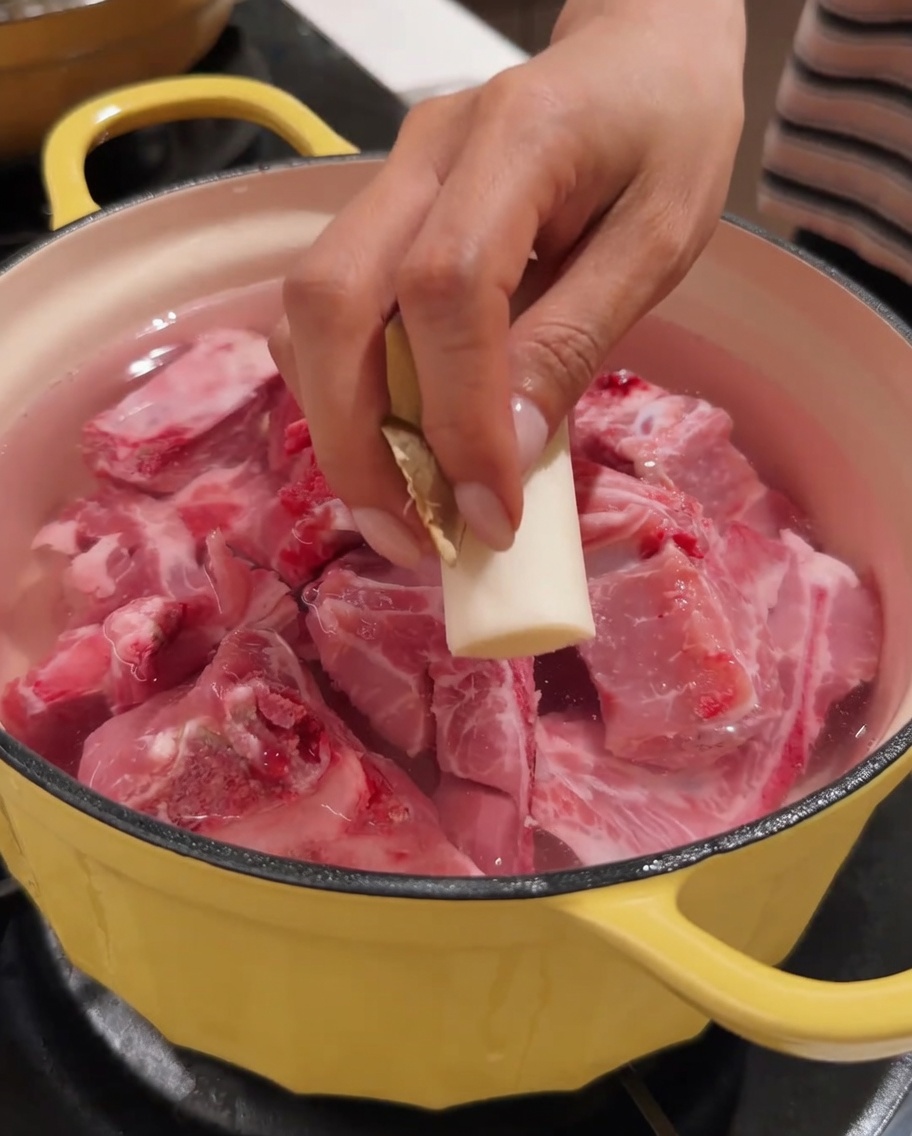 Une main ajoutant un morceau de gingembre dans une marmite contenant du porc et de leau.