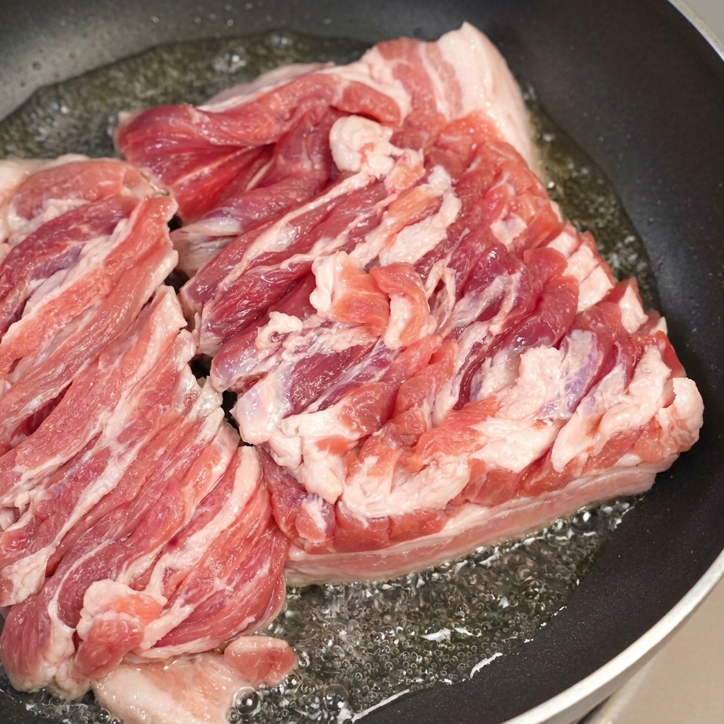 Une poêle avec de la poitrine de porc grésillante et une cuillère retirant lexcès de gras fondu.