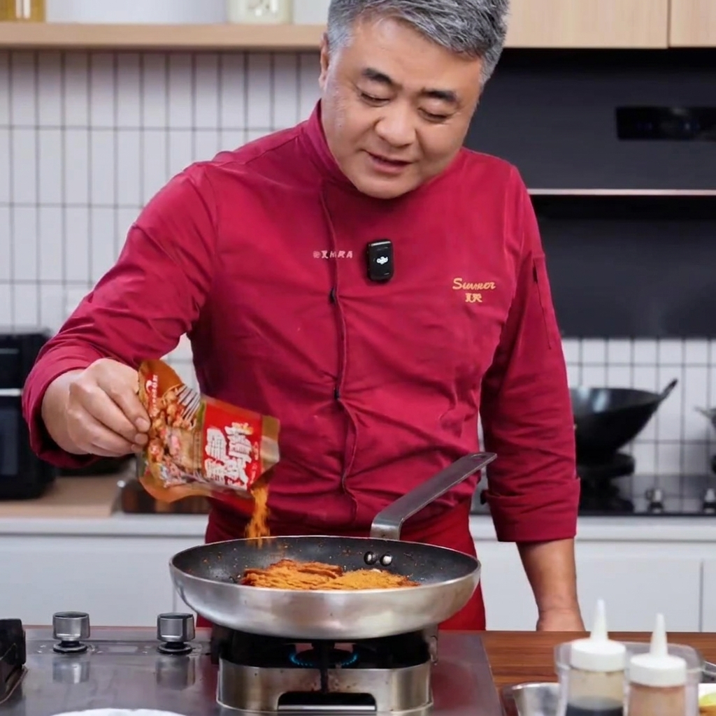 Chef en manteau rouge versant un mélange dépices pour barbecue dun sachet dans une poêle de porc saisi.