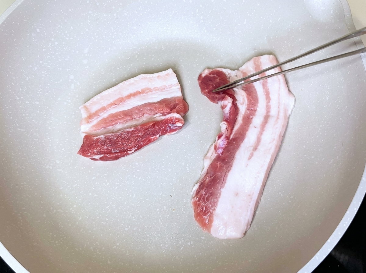 Tranches de poitrine de porc disposées proprement dans une poêle blanche sèche manipulées avec des pinces métalliques.