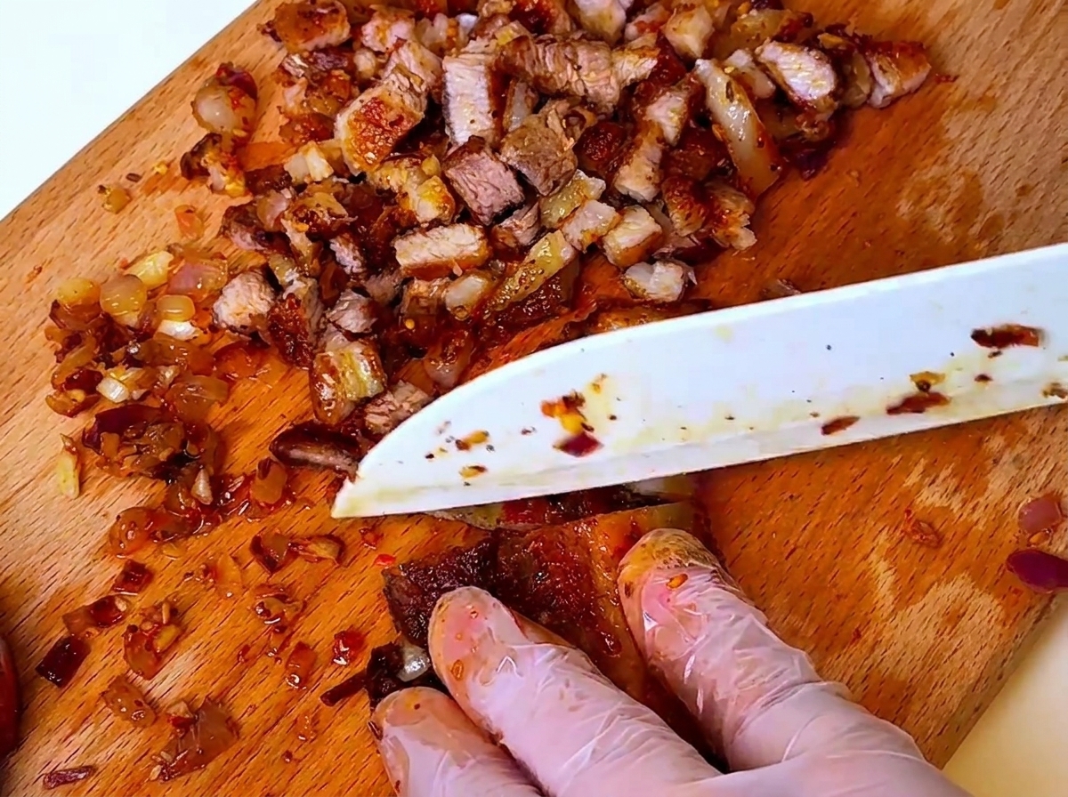 Un couteau coupant du porc barbecue cuit et des oignons sur une planche à découper en bois, avec des mains portant des gants de protection.