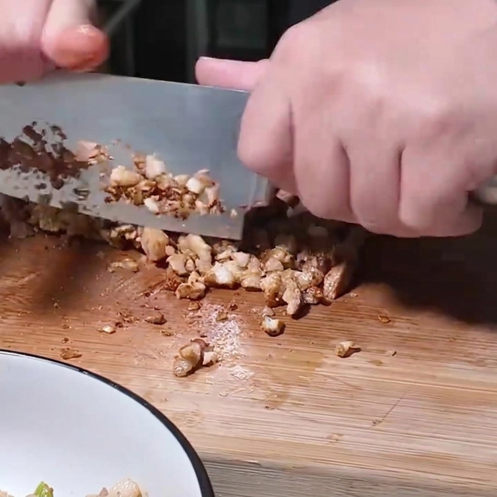 Une personne utilisant un grand couteau pour hacher finement des morceaux de porc cuits sur une planche en bois.
