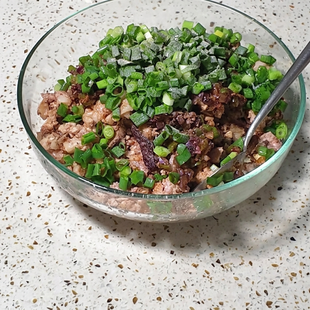 Un bol contenant un mélange de porc et dœufs, garni doignons verts frais et dassaisonnements, en train dêtre remué avec une cuillère.