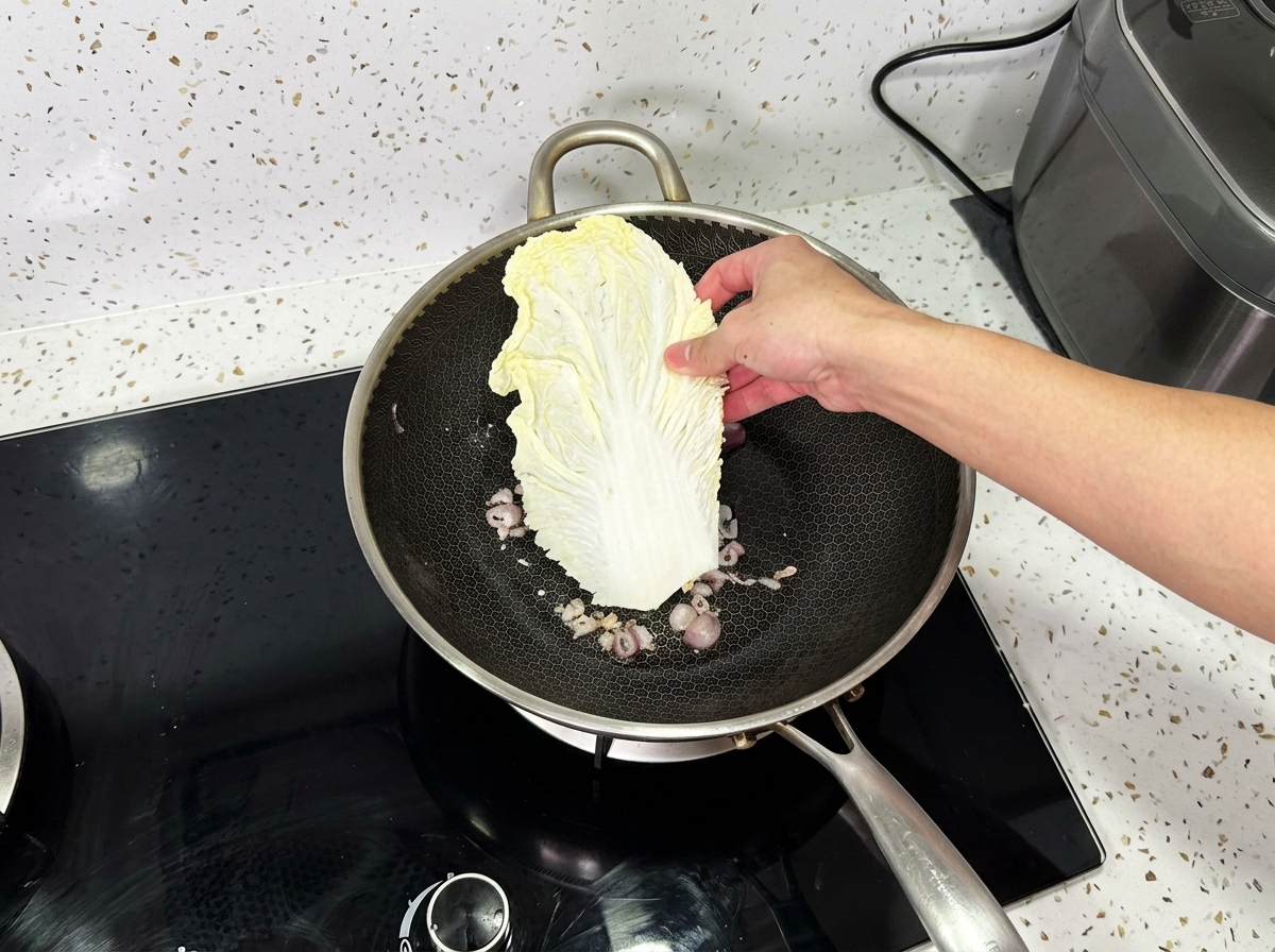 Une main plaçant une grande feuille de chou napa dans un wok contenant des oignons cuits.
