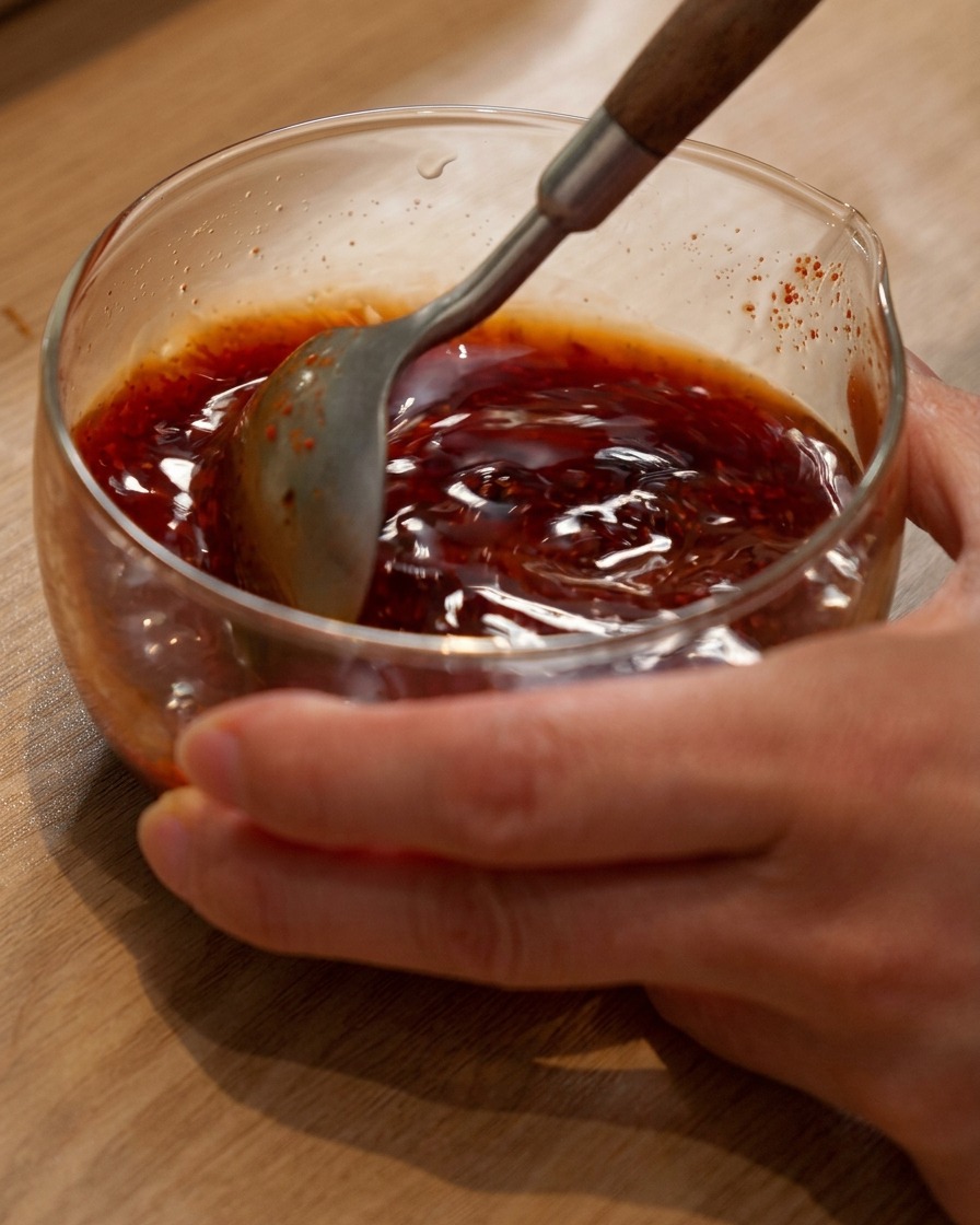 Une cuillère remuant un mélange de sauce brune et rougeâtre pour hot pot dans un bol en verre.