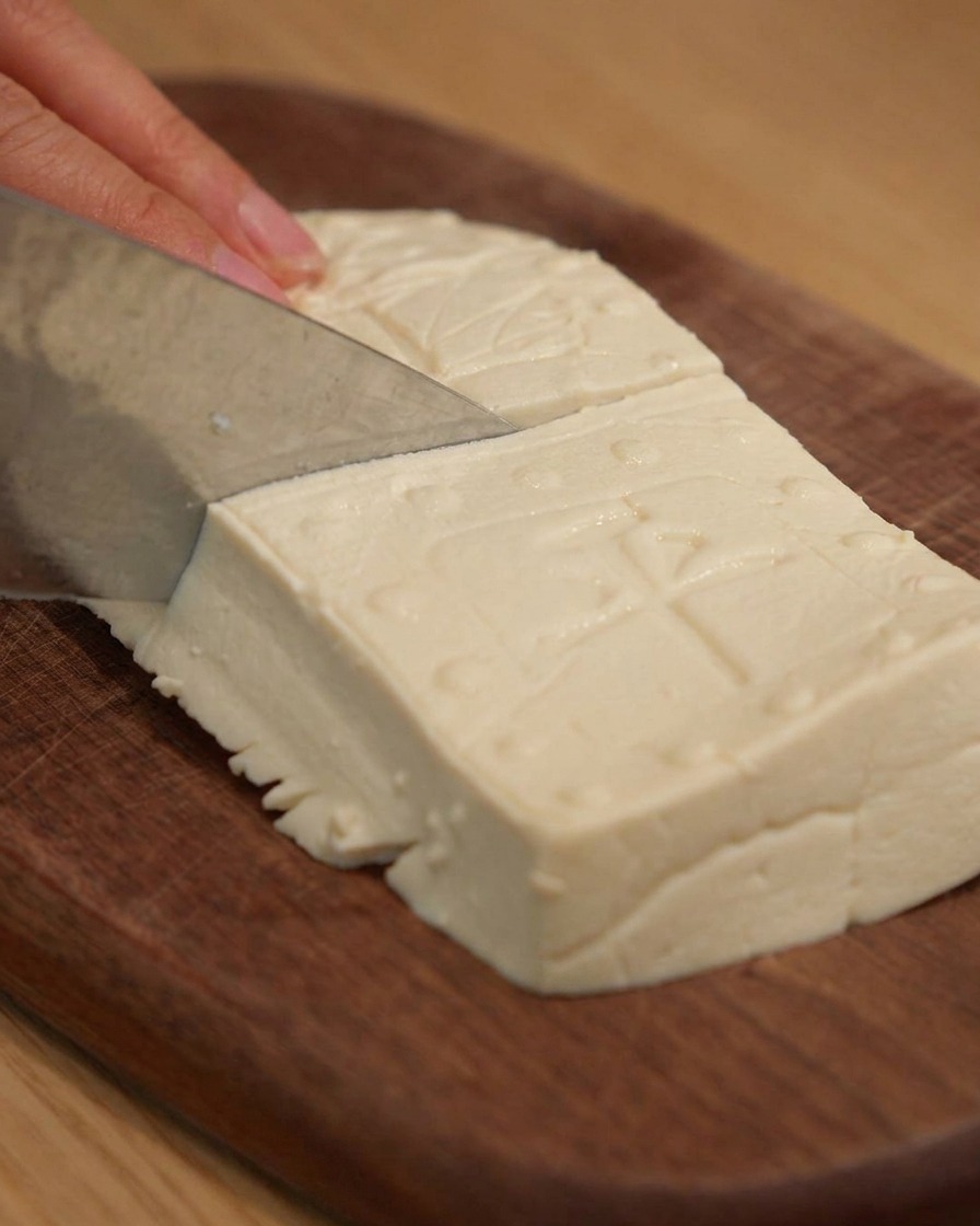 Un couteau bien aiguisé coupant du tofu ferme en blocs rectangulaires nets sur une planche à découper en bois.