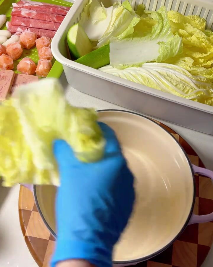 Mains gantées plaçant des feuilles de chou napa dans une casserole mauve.