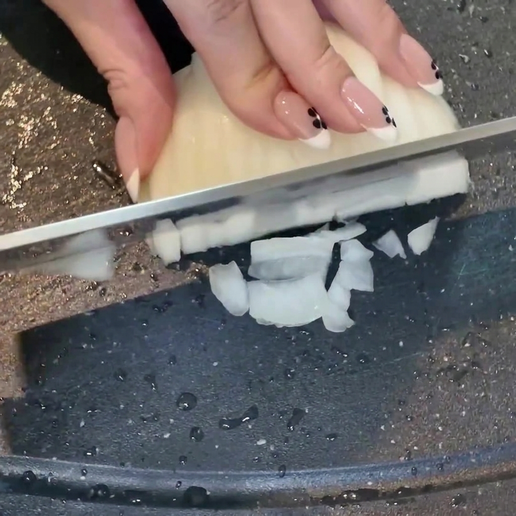 Une main utilisant un couteau pour couper un oignon blanc en dés sur une planche à découper sombre, avec de lail émincé à proximité.
