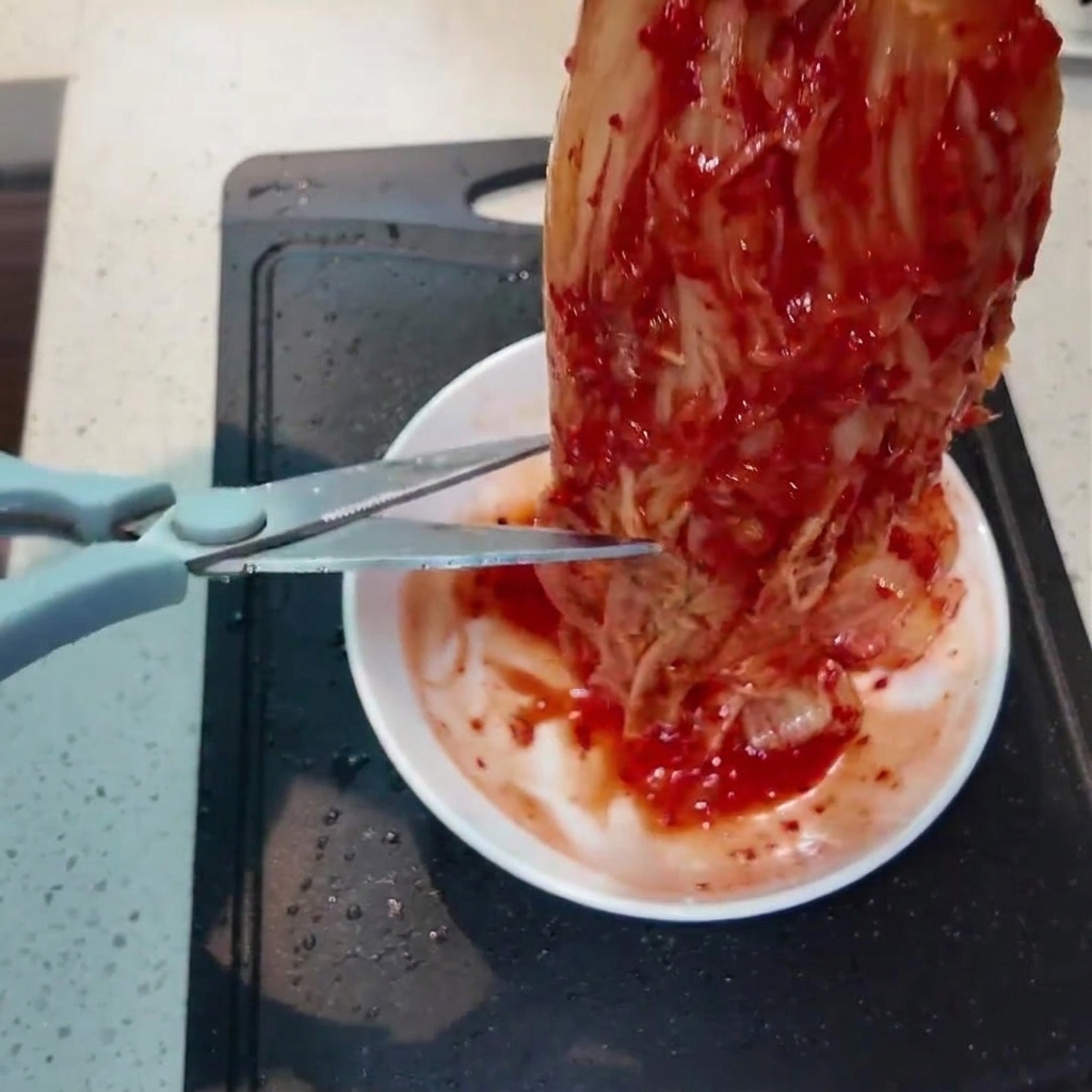 Un morceau de kimchi rouge vif coupé en petits morceaux avec des ciseaux de cuisine au-dessus dun petit plat blanc.