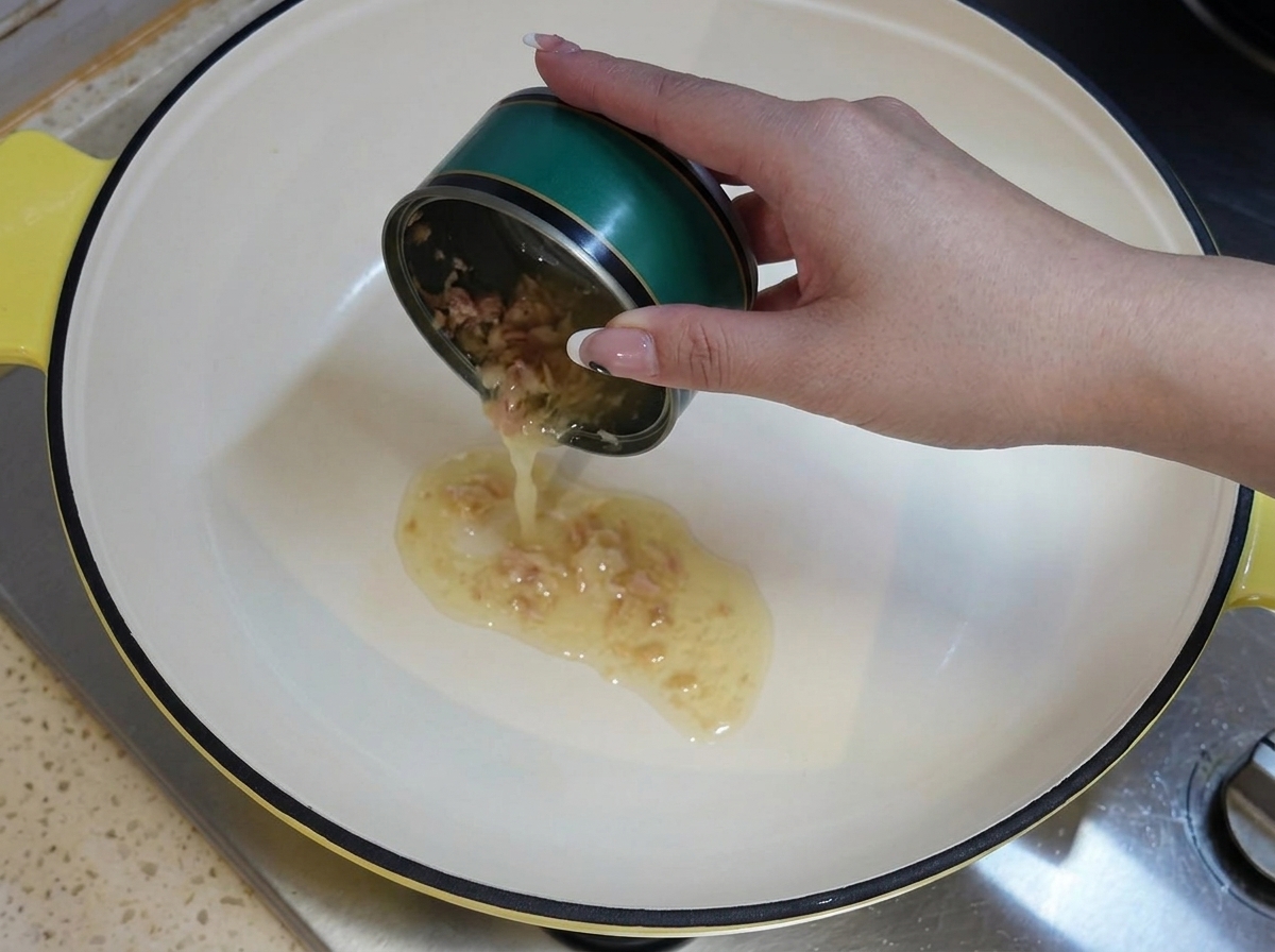 Une main tenant une boîte de thon verte vide, versant son huile résiduelle dans une casserole blanche chauffée sur la cuisinière.
