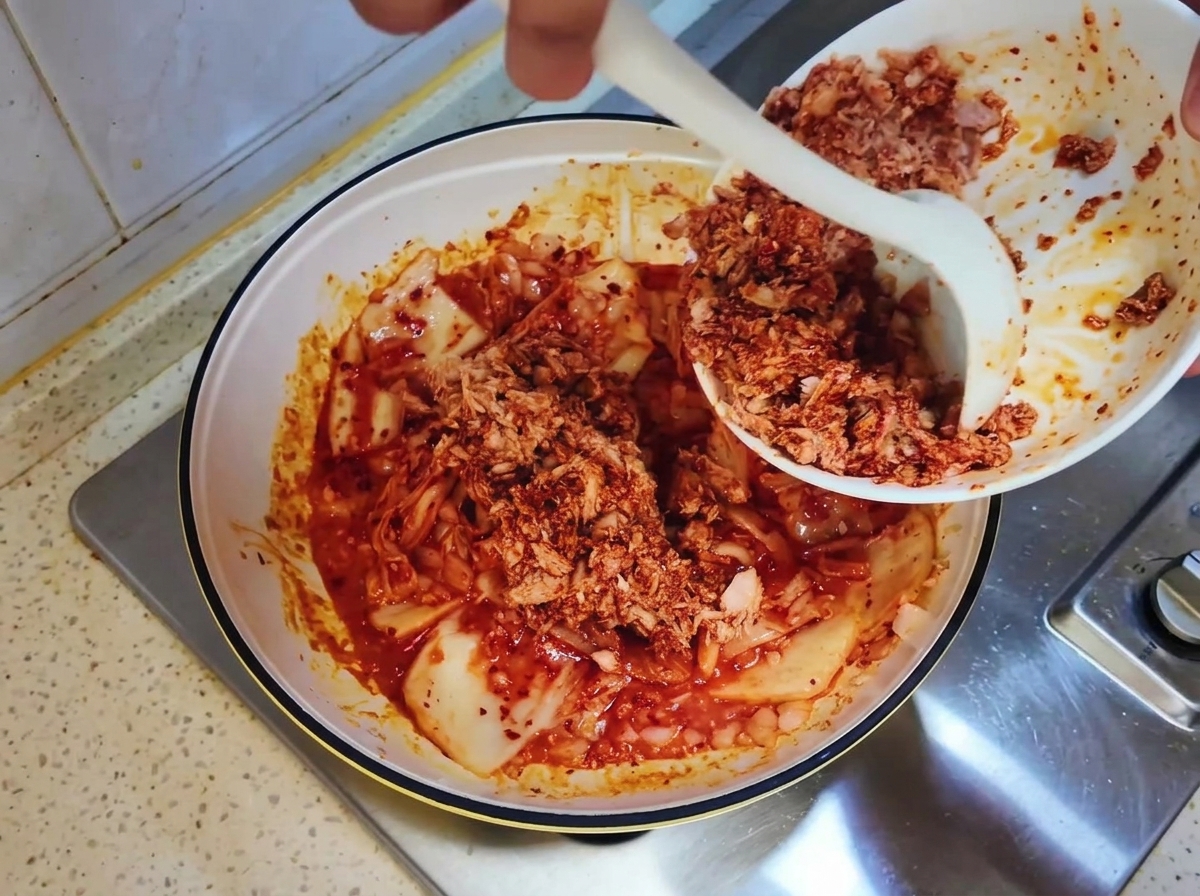 Mélange de thon en conserve raclé dun bol blanc vers la poêle avec le kimchi à laide dune cuillère blanche.