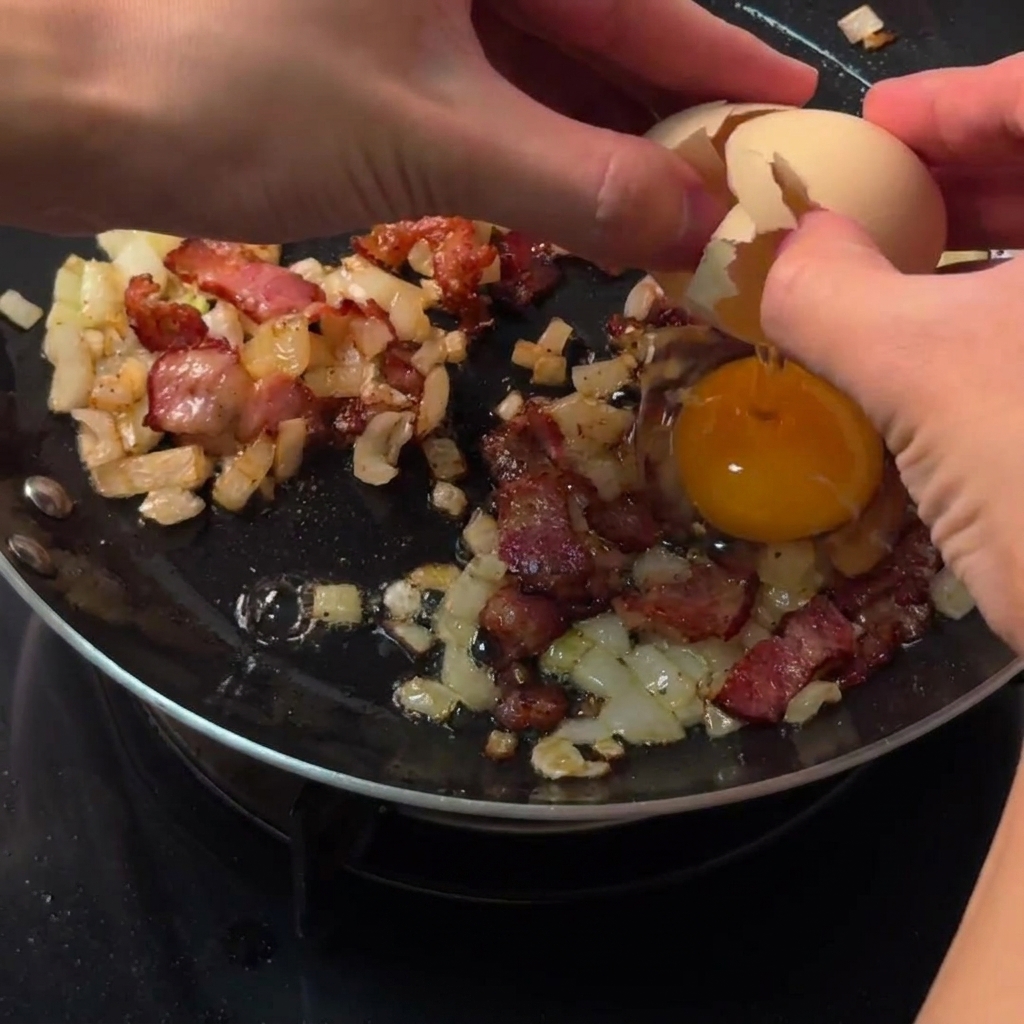 Mains cassant un œuf cru directement dans un espace circulaire dégagé parmi le bacon et les oignons dans une poêle.