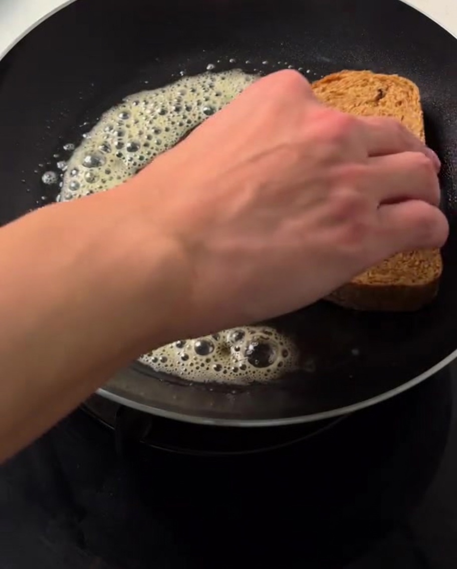 Une main plaçant une tranche de pain complet sur du beurre fondu frémissant dans une poêle chaude.