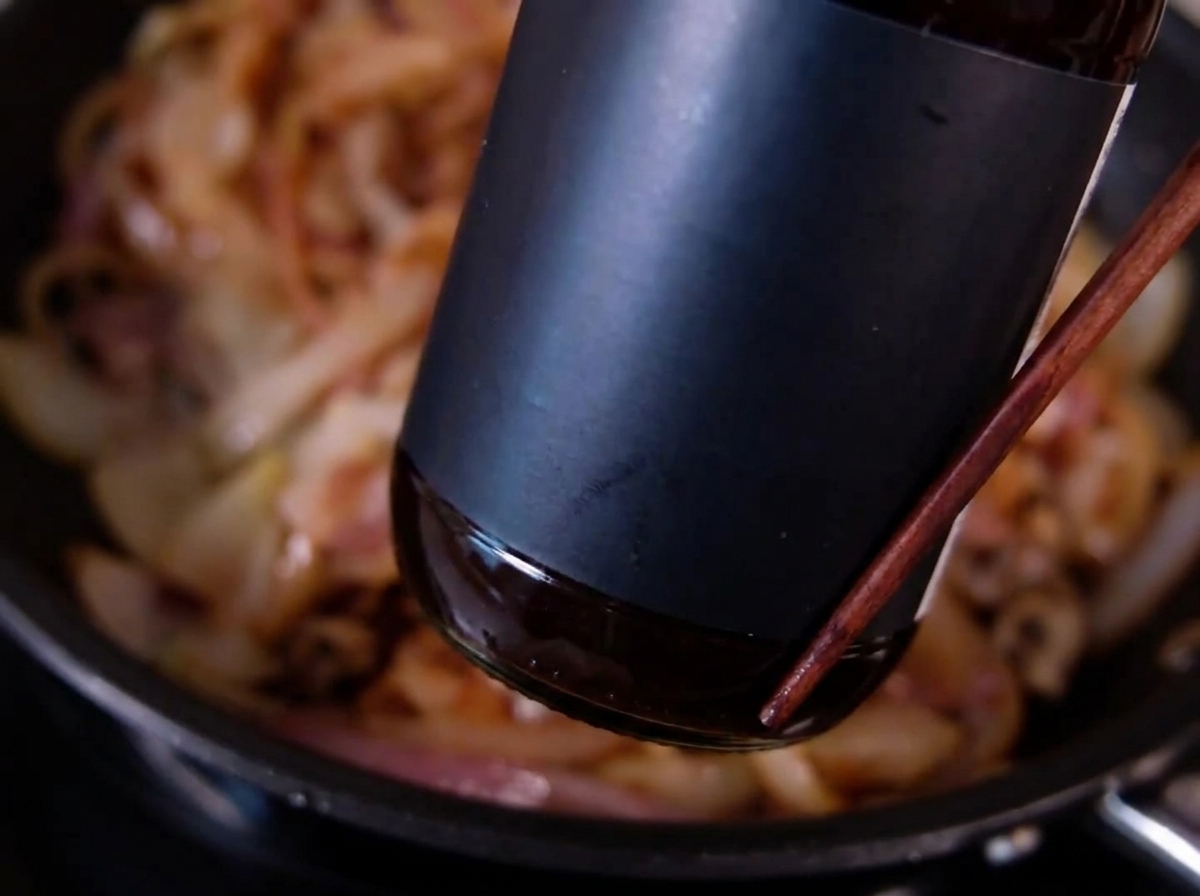 Une bouteille sombre versant de la sauce sur des oignons émincés et dorés cuisant dans une poêle.