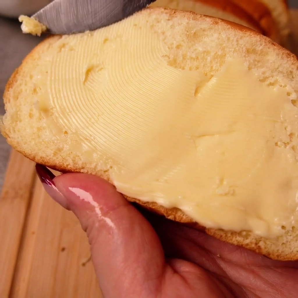 Une main tenant une tranche de pain de mie tout en étalant uniformément du beurre sur sa surface avec un couteau.