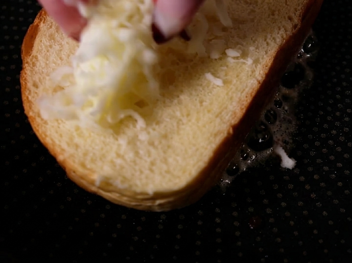 Des doigts déposant de la mozzarella râpée sur une tranche de pain nature dans une poêle chaude.