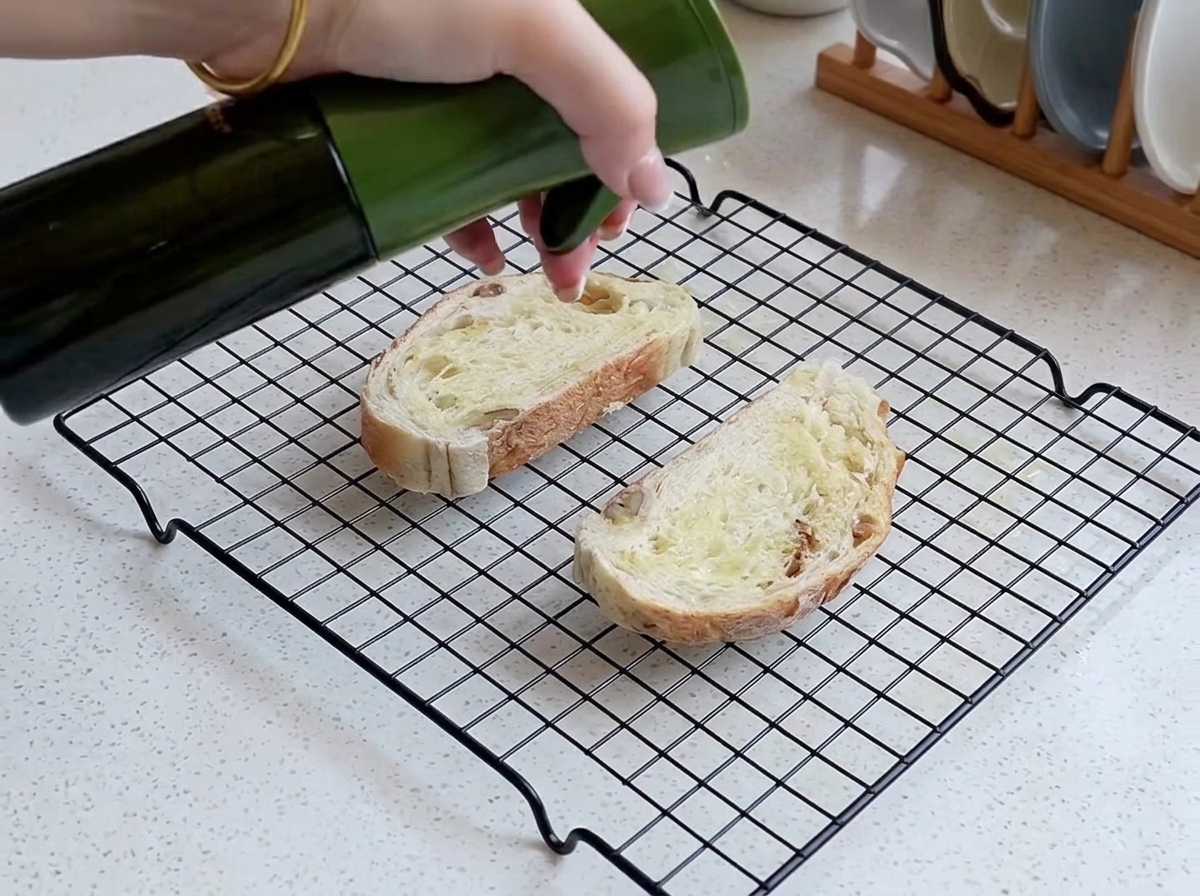 Une main vaporisant une fine couche dhuile sur deux tranches de pain rustique disposées sur une grille métallique.