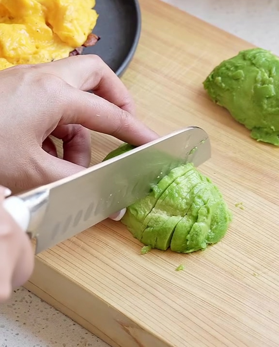 Une main utilisant un couteau de chef pour trancher une moitié davocat épluché en morceaux fins et nets sur une planche en bois.