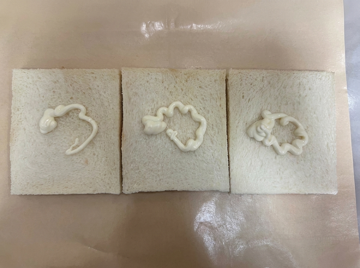 Trois tranches de pain de mie sans croûte alignées sur du papier sulfurisé marron, chacune garnie dun trait ondulé de sauce salade blanchecite: 1.