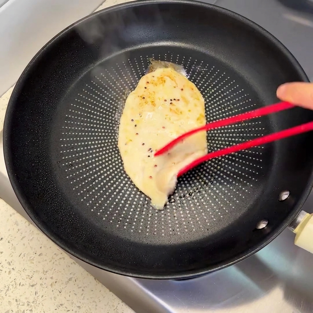 Des pinces rouges retournant une escalope de poulet partiellement cuite dans une poêle, révélant une croûte dorée.