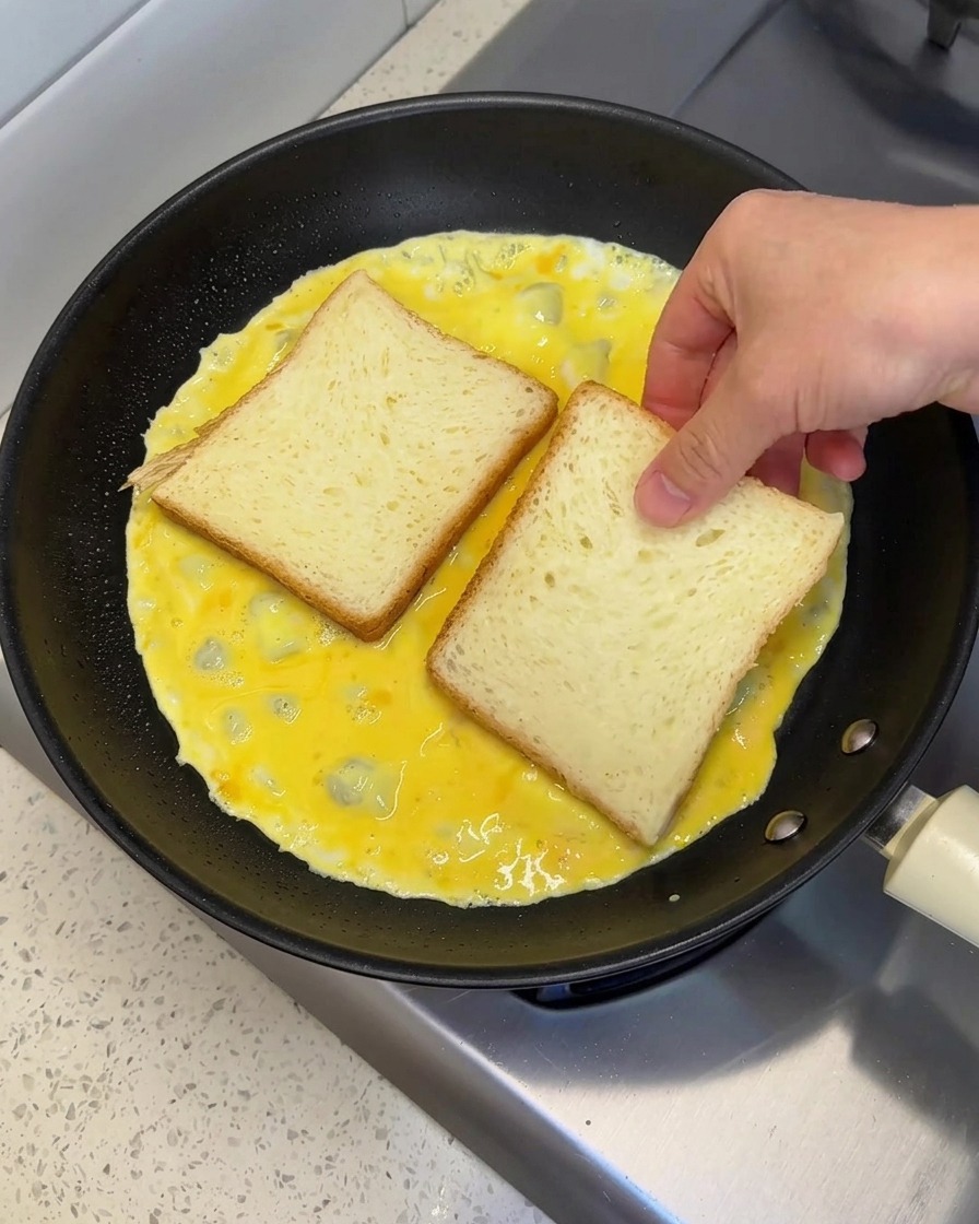 Une main plaçant deux tranches de pain blanc côte à côte sur une omelette aux œufs encore humide dans une poêle.