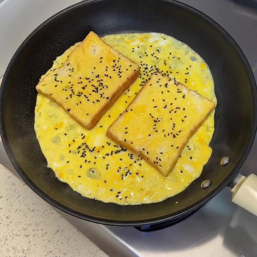 Graines de sésame noir saupoudrées sur deux tranches de pain reposant dans une omelette aux œufs partiellement cuite dans une poêle.
